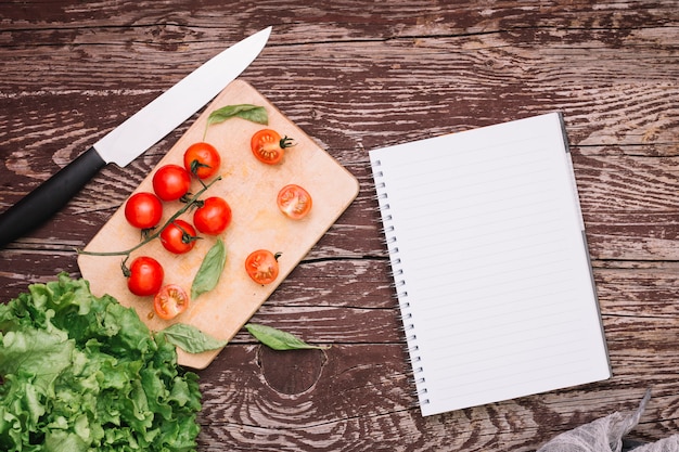 Cuchillo afilado; albahaca; Tomates cherry y lechuga con bloc de notas en espiral sobre superficie de madera