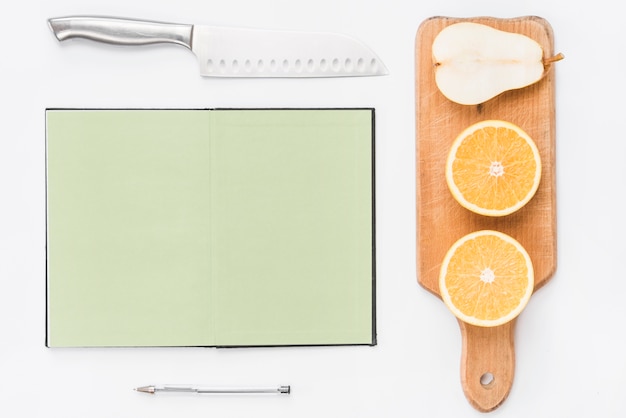 Foto gratuita cuchillo afilado; cuaderno de página en blanco; bolígrafo; mitades de naranjas y peras sobre fondo blanco