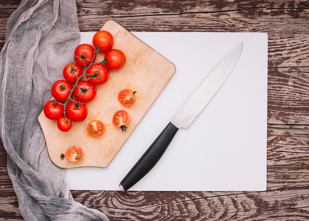 Cuchillo afilado y manojo de tomates cherry en una tabla de cortar sobre el papel blanco contra el escritorio