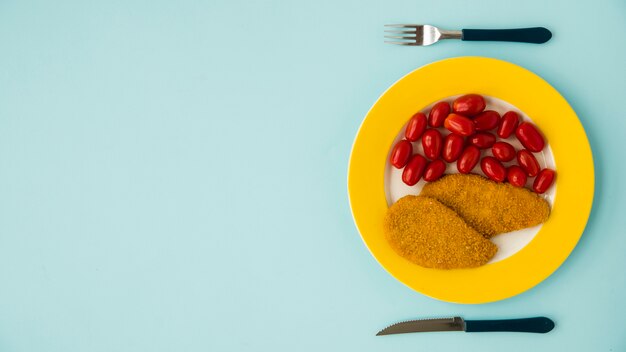 Cuchillo, tenedor y plato con pechuga de pollo y tomate en un escritorio azul
