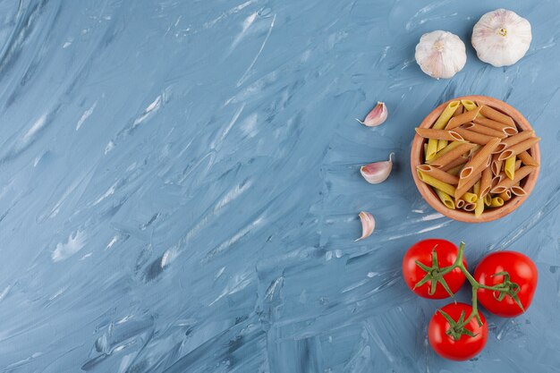 Un cuenco de arcilla de pasta cruda multicolor con ajos y tomates rojos frescos sobre una mesa azul.