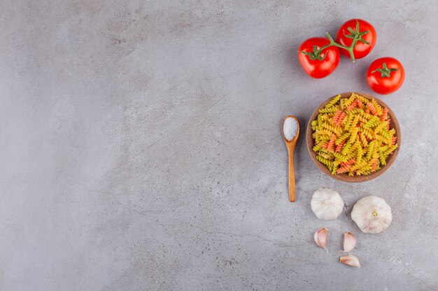 Un cuenco de arcilla de pasta espiral cruda multicolor con ajo y tomates rojos frescos.