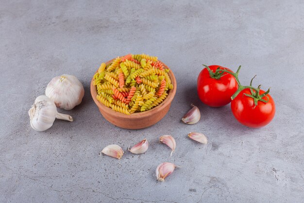 Un cuenco de arcilla de pasta espiral cruda multicolor con ajo y tomates rojos frescos.