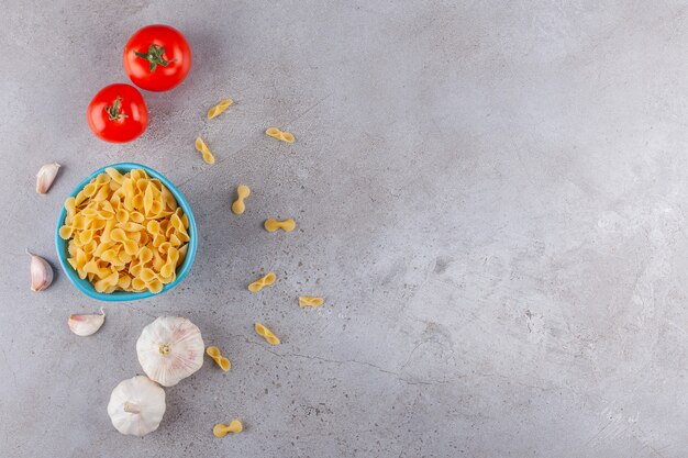 Un cuenco azul lleno de pasta seca cruda Farfalle con verduras sobre una mesa de piedra.