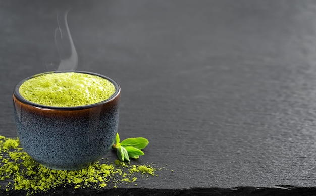 Cuenco de azul con té verde matcha, junto a él hay hojas de té y té en polvo sobre la mesa