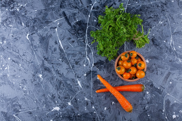 Cuenco de barro de tomates cherry con hojas de perejil y zanahorias sobre fondo azul.