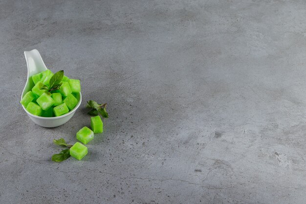 Un cuenco blanco lleno de dulces caramelos verdes con hojas de menta sobre una mesa de piedra.