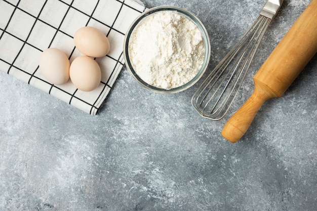 Cuenco de harina, huevos y utensilios de cocina sobre superficie de mármol.