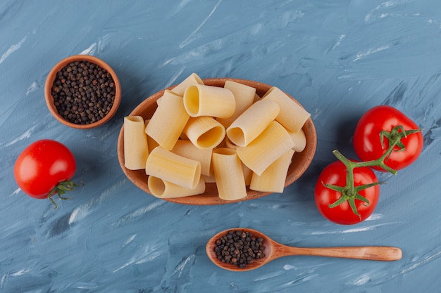 Un cuenco de madera de tubo de pasta seca cruda con tomates rojos frescos y especias sobre una mesa azul.