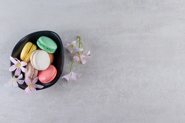 Cuenco negro de coloridos macarrones dulces con flores en la mesa de piedra.