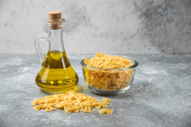 Cuenco de pasta farfalle cruda con botella de aceite de oliva sobre fondo de mármol.