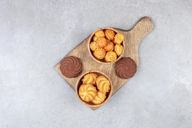 Cuencos de galletas junto a galletas marrones sobre tabla de madera sobre superficie de mármol.