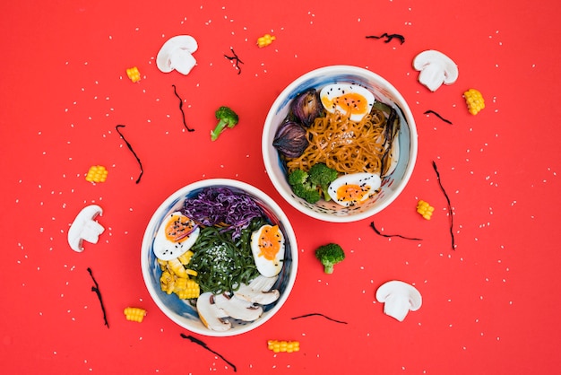 Foto gratuita cuencos ramen picantes con fideos; huevo cocido y verduras servidas con ensalada de algas sobre fondo rojo