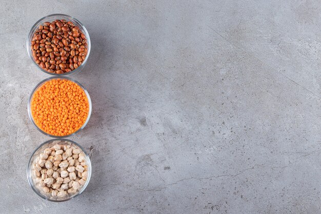 Cuencos de vidrio de frijoles crudos, lentejas y garbanzos sobre fondo de piedra.