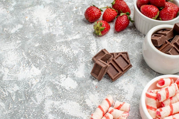 Cuencos de vista de la mitad inferior con fresas, chocolates, dulces y algunas fresas, chocolates, dulces en el lado derecho del fondo de mosaico gris-blanco