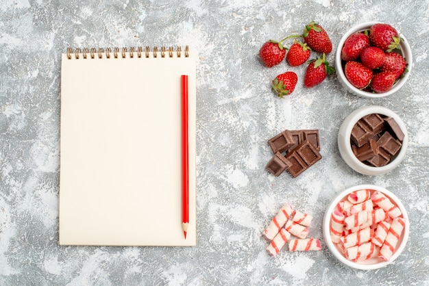 Foto gratuita cuencos de vista superior con fresas, chocolates, caramelos y algunas fresas, bombones, caramelos en el lado derecho y cuaderno con lápiz rojo en el lado izquierdo del suelo gris-blanco.