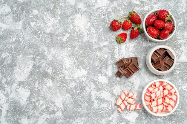 Cuencos de vista superior con fresas, chocolates, caramelos y algunas fresas, bombones, caramelos en el lado derecho del suelo gris-blanco.