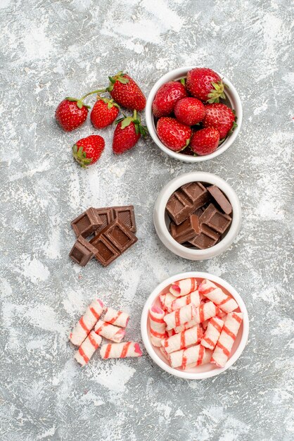 Cuencos de vista superior con fresas chocolates caramelos y algunas fresas chocolates caramelos sobre el suelo gris-blanco