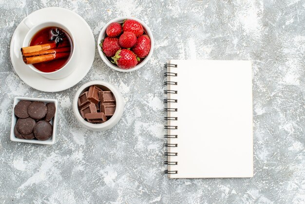 Cuencos de vista superior con fresas y chocolates, té de semillas de anís canela y un cuaderno sobre el suelo gris-blanco