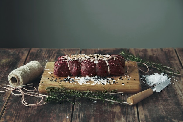 Cuerda atado trozo de carne salada con pimienta lista para fumar en la mesa de madera entre hierbas y especias en madera