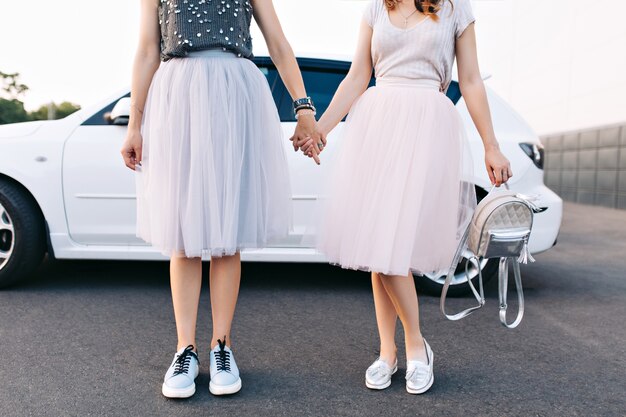 Cuerpos de modelos de moda en faldas de tul y zapatillas de deporte en coche blanco. Se toman de la mano.