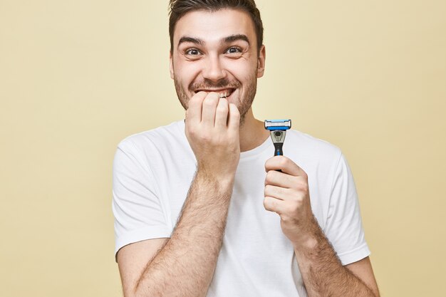 Cuidado de la piel, belleza masculina y concepto de masculinidad. Emocional joven morena con camiseta blanca sosteniendo una navaja de afeitar, con expresión facial nerviosa, mordiéndose las uñas, asustado de afeitarse por primera vez