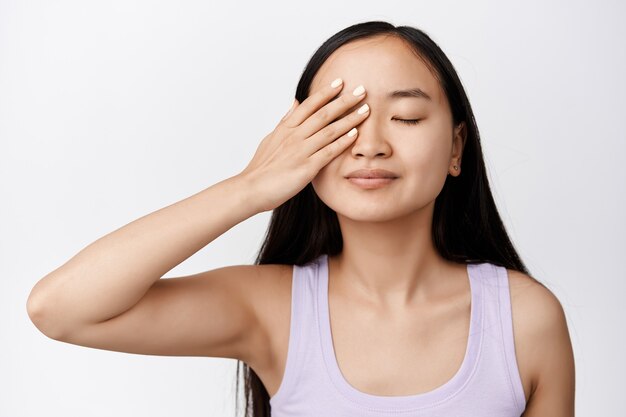 Cuidado de la piel y cosmetología. Hermosa mujer asiática cierra los ojos y sonríe, sosteniendo la mano en la cara, aplicando productos cosméticos para el cuidado de la piel en blanco