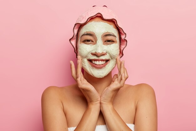 Cuidado de la piel para todas las edades. Feliz dama asiática con mascarilla de arcilla exfoliante en la cara, tiene tratamientos de belleza, se ve agradable, toca las mejillas, usa gorro de ducha