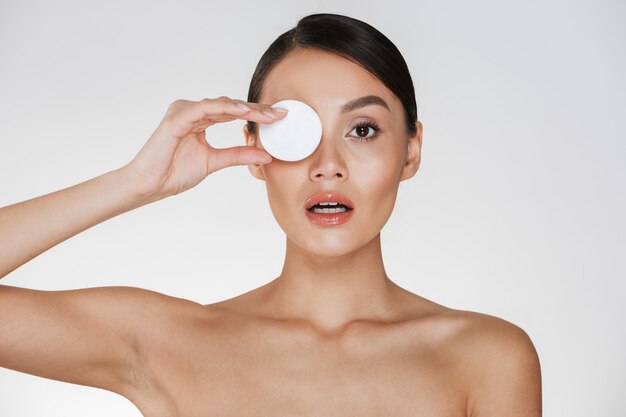 Cuidado de la piel y tratamiento saludable de la mujer que se pone un algodón en el ojo mientras se quita los cosméticos de la cara, aislados en blanco