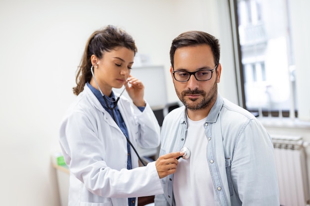 Cuidadosa doctora caucásica usa fonendoscopio examina la frecuencia cardíaca del paciente masculino en la consulta en el hospital Mujer enfermera o médico de cabecera usa estetoscopio escucha los latidos del corazón del hombre en la clínica