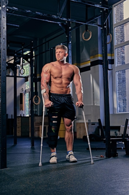 Un culturista sin camisa con muletas cerca de un soporte cruzado en un gimnasio.