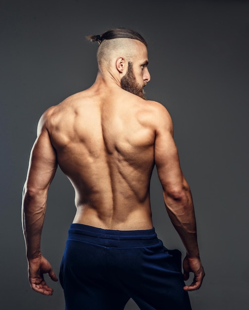 Culturista musculoso sin camisa con barba posando sobre fondo gris.