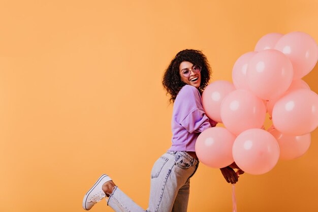 Cumpleañera de buen humor en jeans posando sobre fondo naranja Encantadora dama negra bailando con globos de fiesta