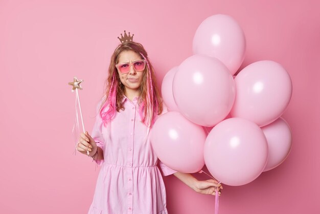 La cumpleañera disgustada e irritada tiene la imagen de la princesa con gafas de sol de corona y el vestido sostiene una varita mágica y un montón de globos en una fiesta aislada sobre un fondo rosa. Concepto de celebración