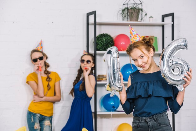 Foto gratuita una cumpleañera que muestra el número 16 de globos plateados con sus dos amigas soplando la fiesta