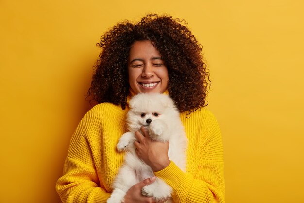 cumpleañera sonríe ampliamente, obtiene una mascota encantadora como regalo, sueña con tener spitz mucho tiempo, usa un jersey amarillo, se para en el interior