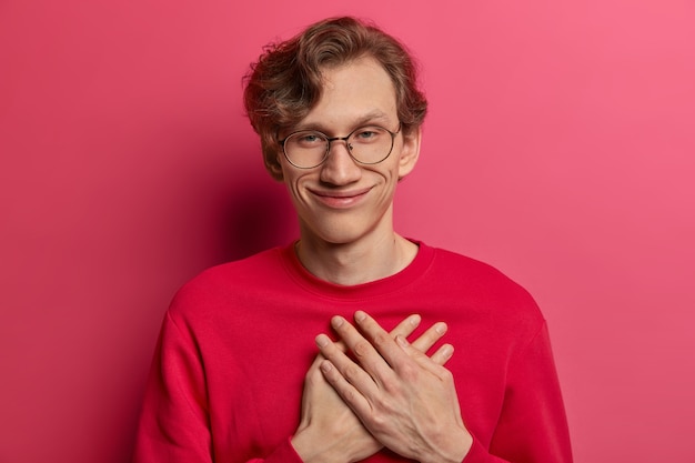 El cumpleañero sonriente y complacido conmovido hace un gesto de agradecimiento, mantiene las palmas de las manos presionadas en el corazón, expresa gratitud por las conmovedoras felicitaciones, gracias por los regalos, usa un jersey rojo informal