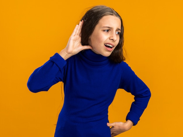 Foto gratuita curiosa adolescente mirando a un lado tocando la cintura haciendo no puedo oírte gesto aislado en la pared naranja