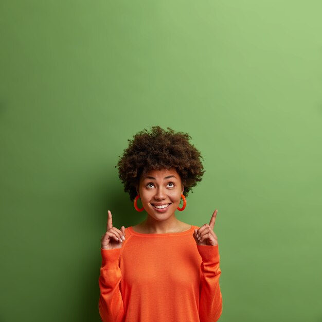 Curiosa y complacida modelo femenina de piel oscura mira hacia arriba y señala el espacio vacío, demuestra la promoción, sugiere subir las escaleras, aislado en la pared verde. Ver esta oferta