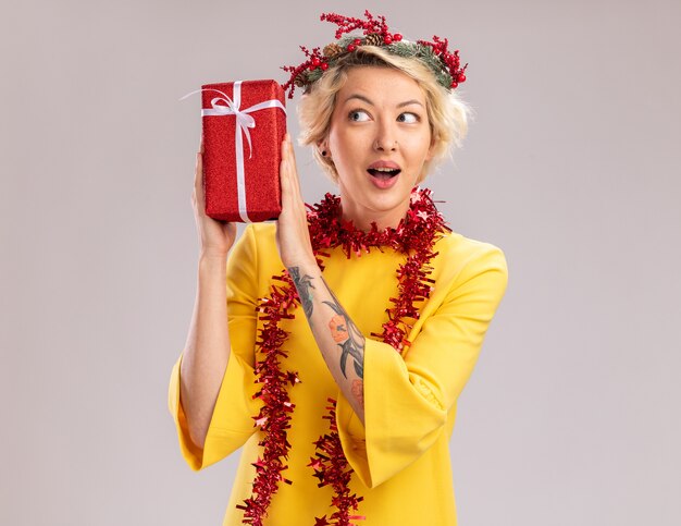 Curiosa joven mujer rubia con corona de Navidad y guirnalda de oropel alrededor del cuello sosteniendo el paquete de regalo de Navidad cerca de la cabeza mirándolo aislado sobre fondo blanco.