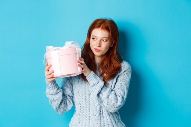 Curiosa jovencita con el pelo rojo, agitando la caja de regalo y preguntarse qué hay dentro, de pie sobre fondo azul.