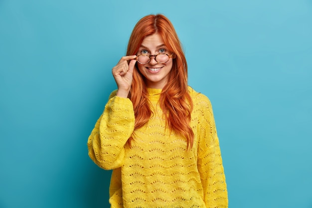 Curiosa mujer europea alegre mira a través de gafas transparentes viste un suéter amarillo de punto sonríe alegremente.