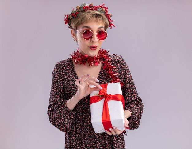Curiosa niña bonita con corona de Navidad y guirnalda de oropel alrededor del cuello con gafas sosteniendo el paquete de regalo agarrando la cinta aislada en la pared blanca con espacio de copia