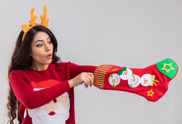 Curiosa niña bonita con diadema de cuernos de reno y suéter de santa claus sosteniendo y mirando la media de Navidad poniendo la mano dentro aislado sobre fondo blanco.