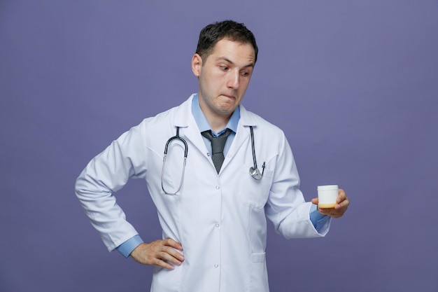 Curioso joven médico masculino con bata médica y estetoscopio alrededor del cuello manteniendo la mano en la cintura sosteniendo un contenedor de tabletas mirándolo aislado en un fondo morado