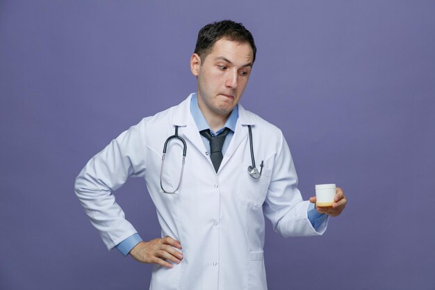 Curioso joven médico masculino con bata médica y estetoscopio alrededor del cuello manteniendo la mano en la cintura sosteniendo un contenedor de tabletas mirándolo aislado en un fondo morado