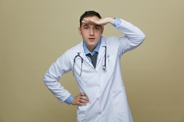 Curioso joven médico masculino con túnica médica y estetoscopio alrededor del cuello manteniendo las manos en la cintura y en la frente mirando la cámara a distancia aislada en un fondo verde oliva