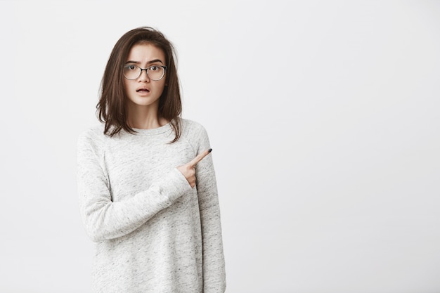 Cutie joven con gafas y sudadera blanca apuntando a un lado con expresión de asombro y confusión.