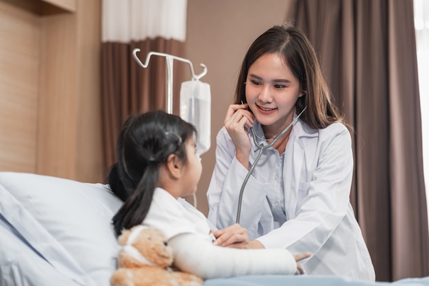 D asiático = médico pediatra examinando a una niña en la cama con un osito de peluche con un estetoscopio