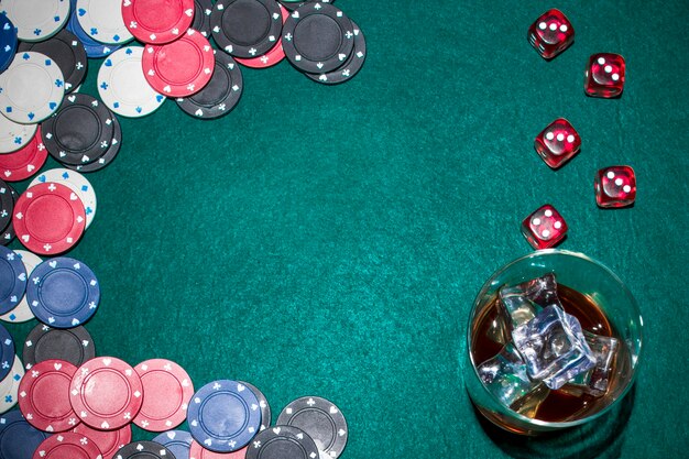 Dados rojos; fichas de casino y vaso de whisky con cubitos de hielo en la mesa de póquer verde